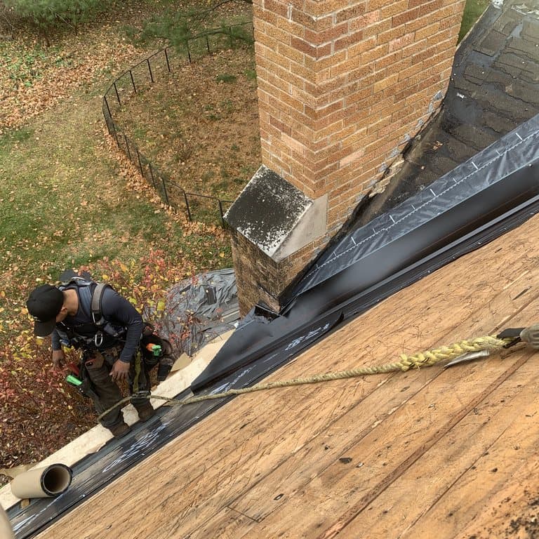 Roofer in a harness detailing ice and water shield around a brick chimney
